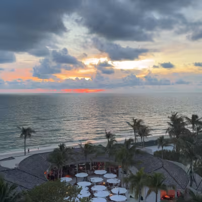 Dramatic Gulf of Mexico sunset with glowing orange and pink light beneath dark clouds, overlooking a beachfront resort terrace filled with white umbrellas and palm trees