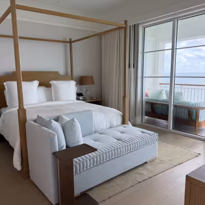 Elegant oceanfront hotel bedroom with a wooden four-poster bed, pale blue upholstered bench, and sliding doors opening to a balcony with a striped outdoor daybed and sea views
