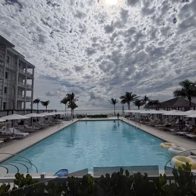 Expansive resort pool framed by lounge chairs and white umbrellas, with palm trees and the ocean in the distance under a dramatic, cloud-filled sky