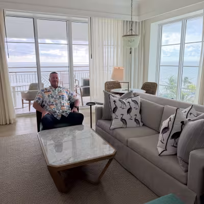 Bright, coastal-style hotel living room with large windows and balcony views of the ocean, a gray sofa with bird-print pillows, a marble coffee table, and a man seated in an armchair.