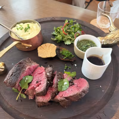 Sliced medium-rare Chateaubriand steak on a wooden board with mashed potatoes in a copper pot, chimichurri, jus, roasted garlic, and a small green salad