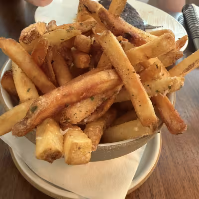 Bowl of golden, thick-cut French fries piled high on a restaurant table, with a diner in a white top holding a fry in the background