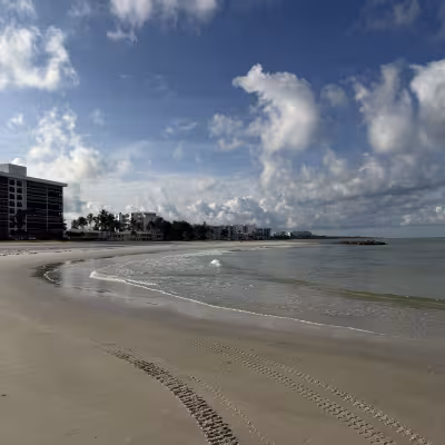 Wide sandy beach with gentle waves, tire tracks in the sand, and a line of beachfront buildings in the distance under a bright blue sky with scattered clouds