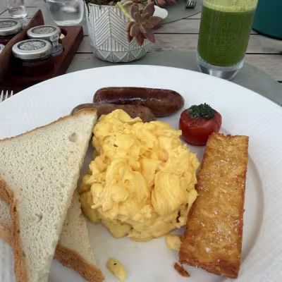 Hotel breakfast plate with scrambled eggs, sausage links, toast, and a crispy hash brown, served outdoors with a green juice and small potted succulent on the table