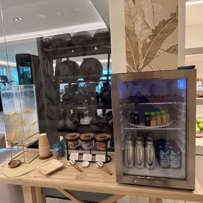 Fitness center refreshment station with infused water, jars of healthy snacks, and a mini fridge stocked with juices, wellness shots, and coconut water next to a glass wall of gym equipment