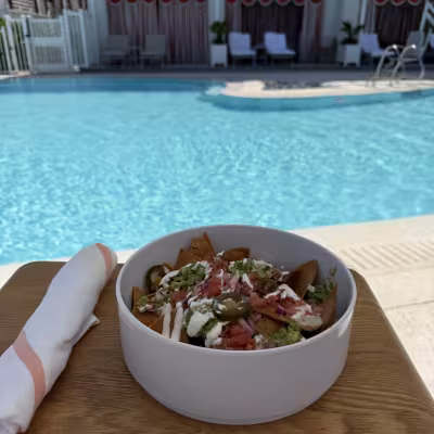 Bowl of loaded nachos topped with guacamole, jalapeños, tomatoes, onions, and crema on a wooden side table beside a bright blue resort pool and private cabanas
