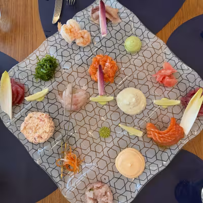 Artful seafood platter on a patterned glass plate with assorted sashimi, tartares, sauces, and garnishes arranged in a circle on a wooden restaurant table