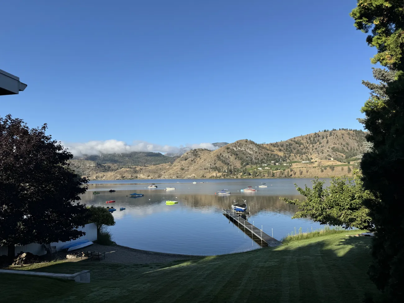 Calm Okanagan lakefront with small boats anchored below golden hills on a clear morning