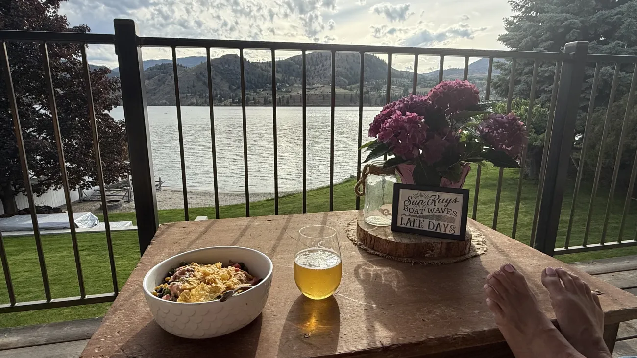 Relaxed lakefront balcony scene with a meal, drink, flowers, and mountain views