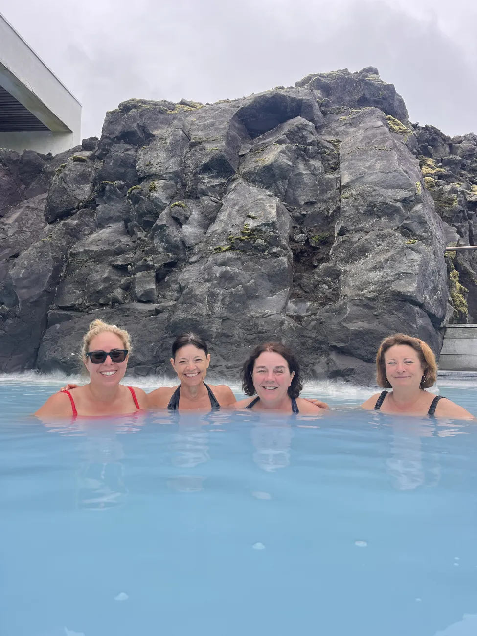 Four women smiling and relaxing in milky blue geothermal water, backed by dark volcanic rock walls at a spa pool.
