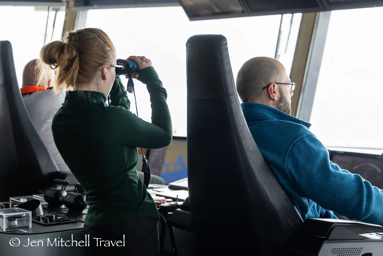 Expedition bridge scene with a woman using binoculars beside crew at the helm, scanning the bright horizon through large windows.