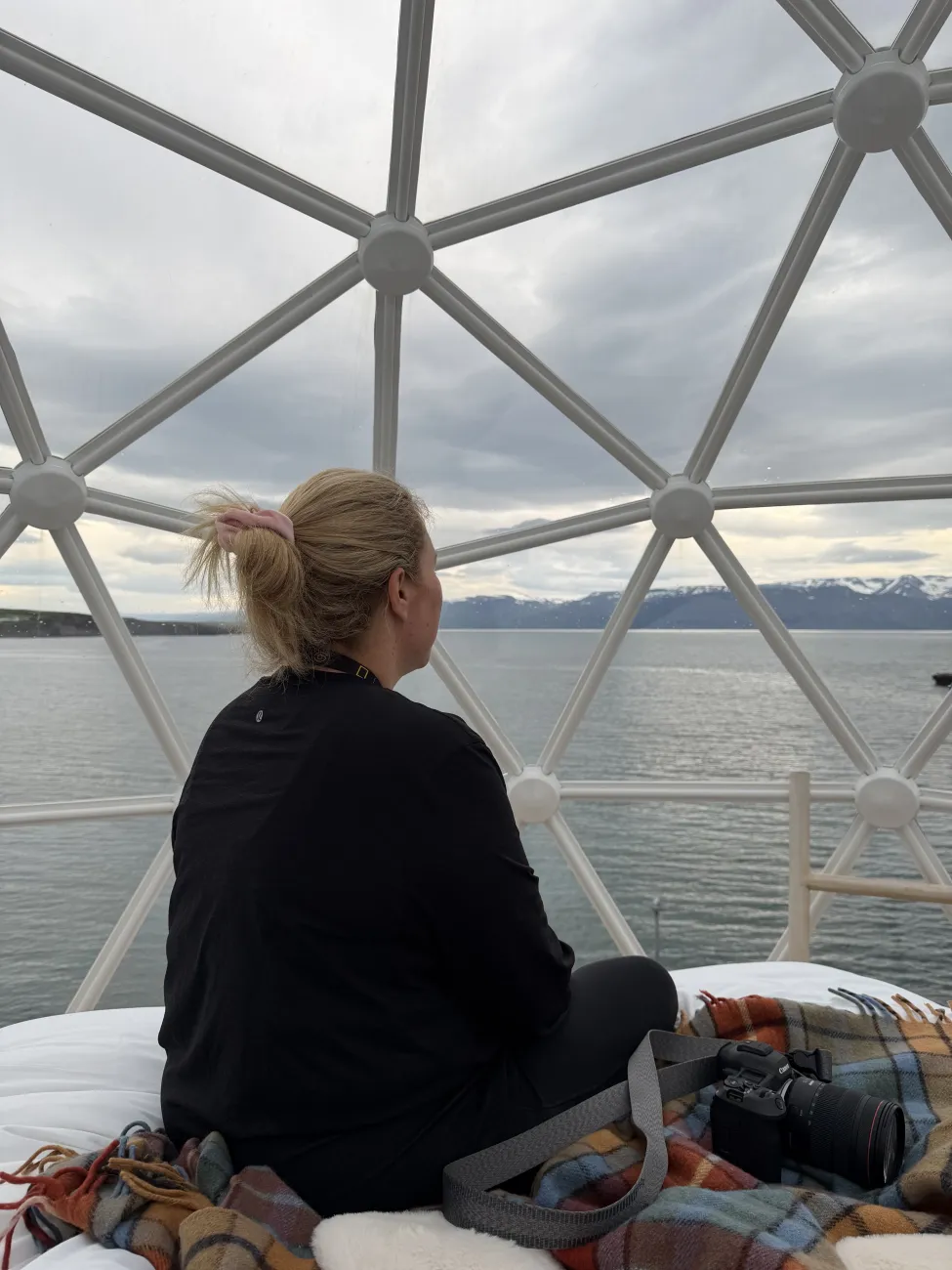 Woman sitting inside a glass igloo-like dome, gazing out over calm water and distant snow-capped mountains with a camera resting on a blanket.