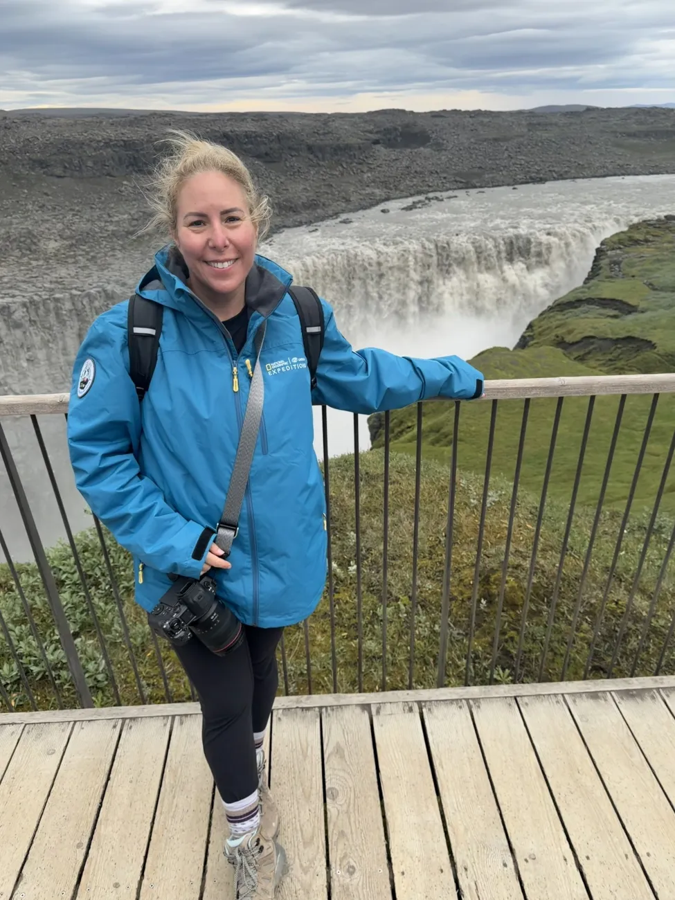 Smiling woman in a blue expedition jacket standing at a viewpoint above a wide, powerful waterfall cutting through a rocky green canyon.