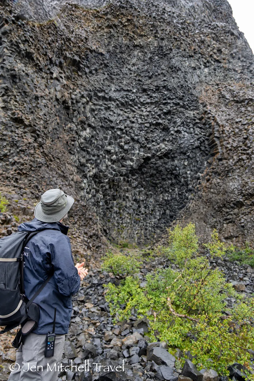 Expedition guide in rain gear and backpack standing at the base of a towering basalt rock wall, observing the geometric columns.