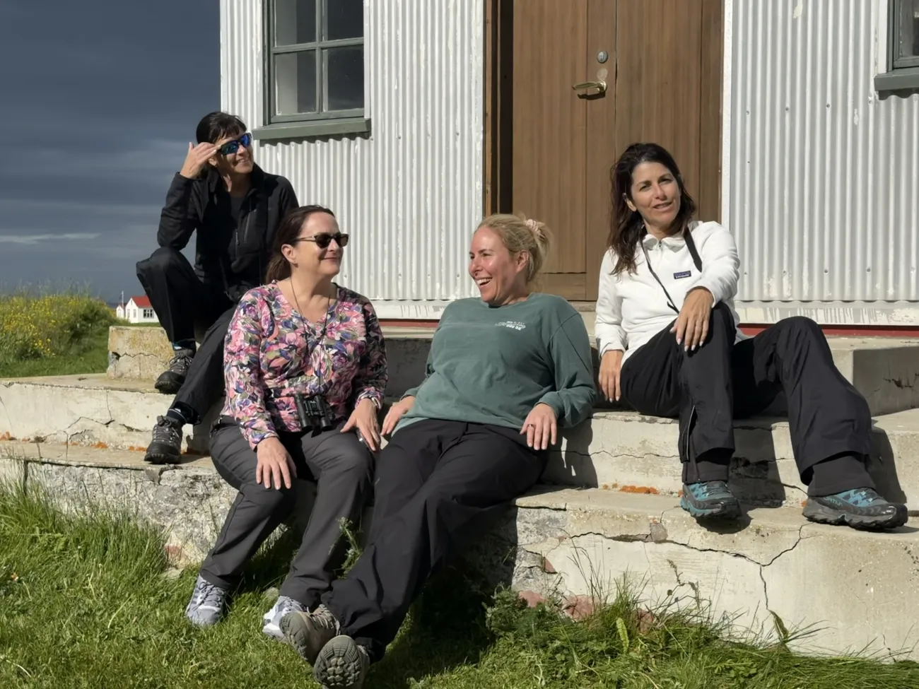 Four women sitting on the sunlit steps of a simple building, laughing and relaxing in outdoor gear with a stormy sky and yellow flowers in the background.