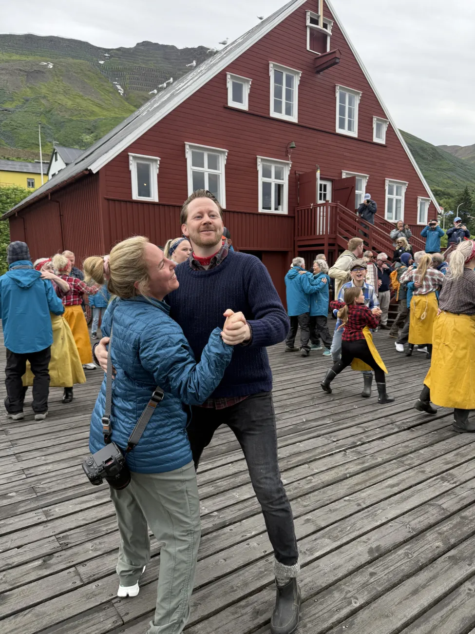 Woman dancing and laughing with a local man on a wooden dock in front of a red historic building during a lively herring museum performance.