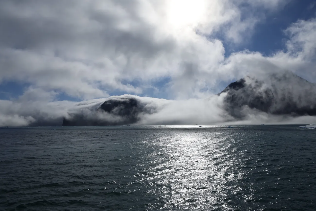 Sunlight glints off a calm Arctic bay as low clouds and fog wrap around dark, rugged mountains rising from the water.